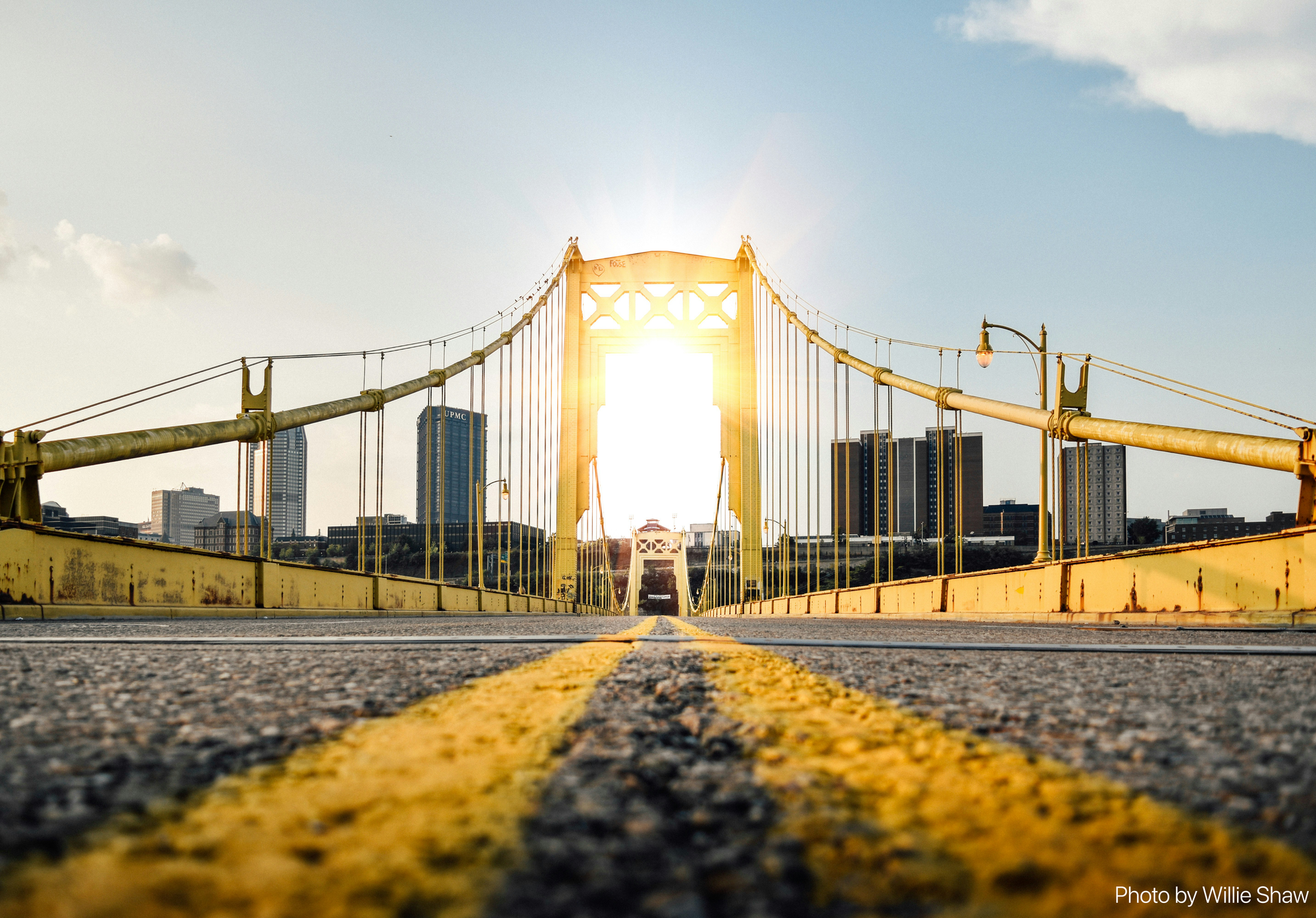 A artistic shot of one of the Three Sisters bridges where the amera is sitting on the ground between the two yellow lines of the road, facing the towers of the cable bridge, with the skyline and sun behind.